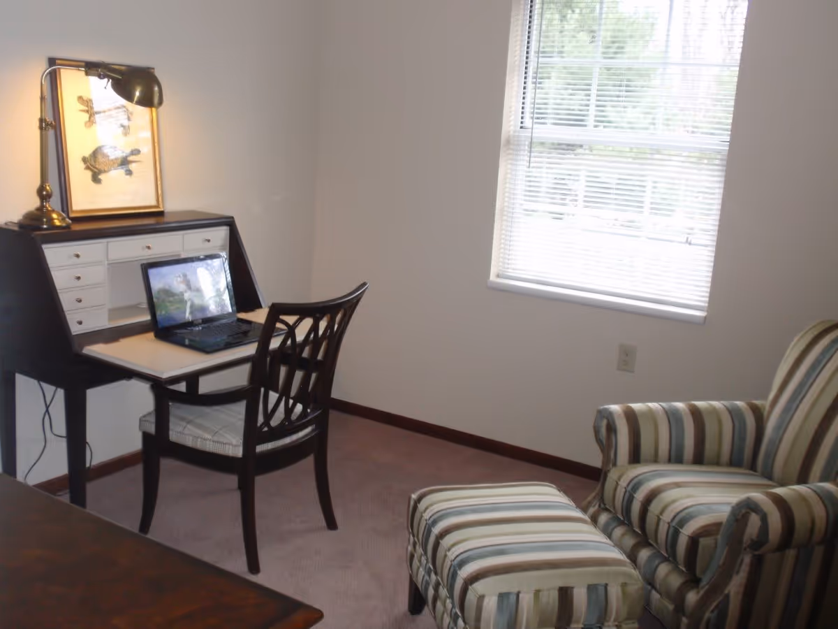 A small room with a window covered by blinds, a striped upholstered armchair with a matching ottoman, and a dark wood desk with a chair. On the desk is a laptop and a brass desk lamp illuminating a framed picture of a turtle.