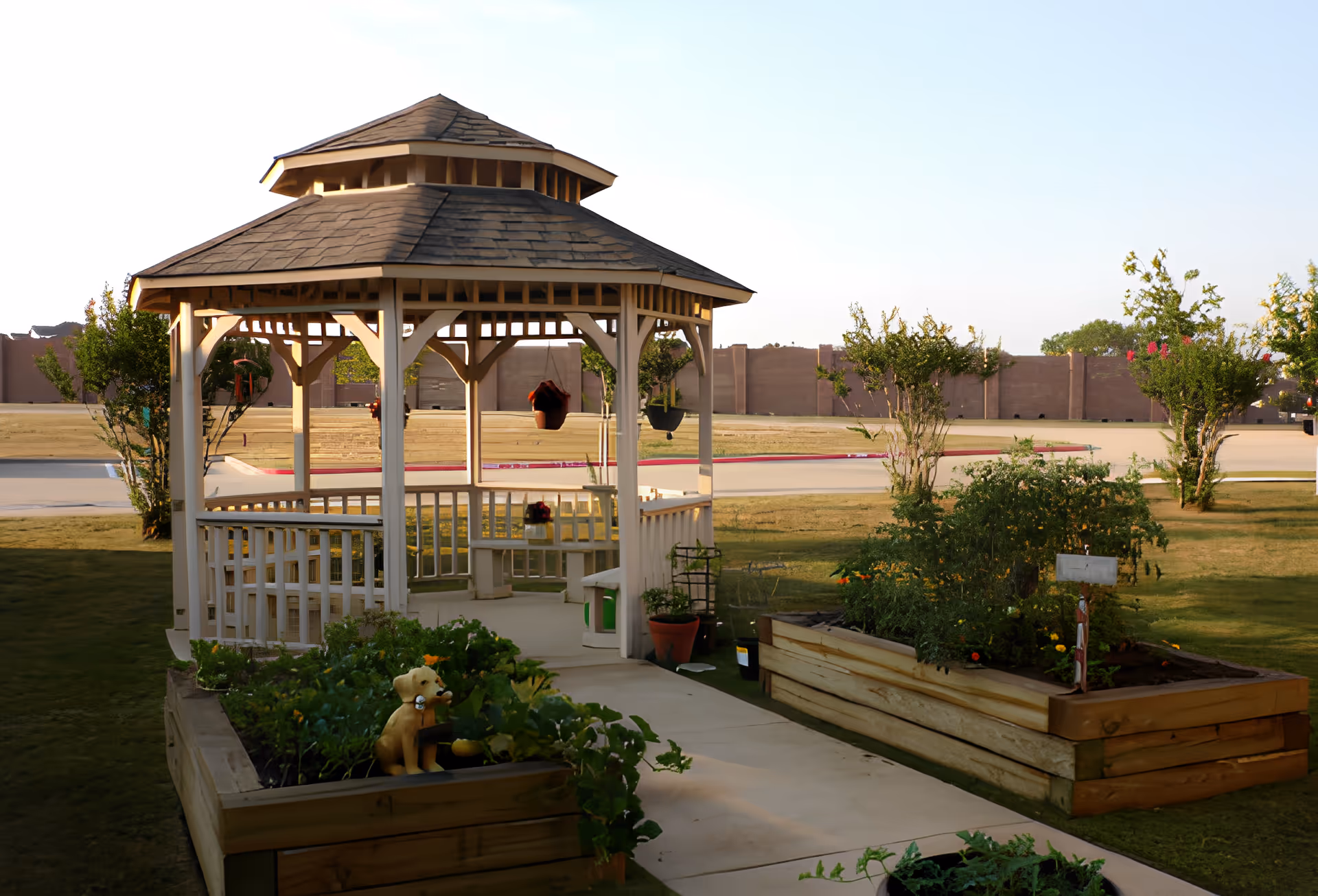 A white wooden gazebo on a lawn surrounded by raised garden beds and potted plants.