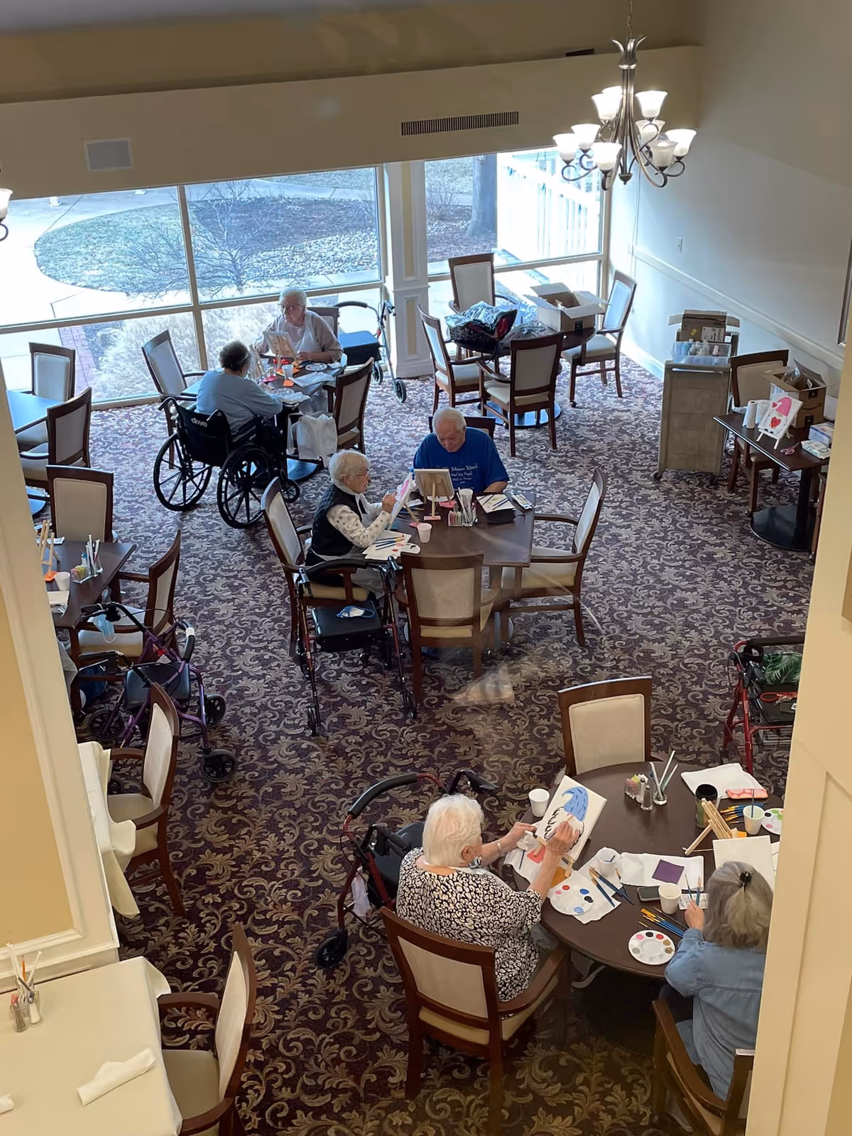 A group of elderly people sitting around tables in a well-lit room with large windows, engaging in painting and other arts and crafts activities. Some individuals are in wheelchairs or using walkers. The room has patterned carpet and several chairs and tables arranged for group activities.