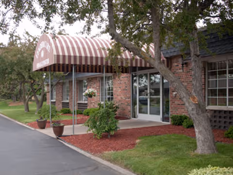 Brick building entrance with a striped awning, glass doors, potted plants, and landscaping under a tree.