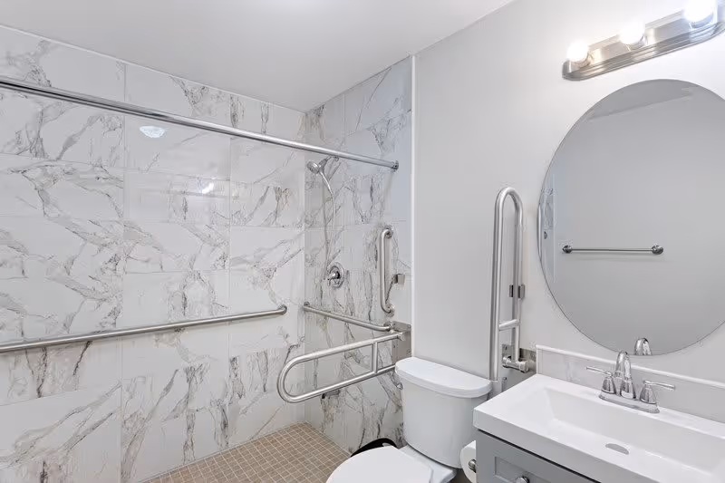 A modern bathroom with marble-patterned wall tiles and beige floor tiles. It features a walk-in shower with multiple stainless steel grab bars, a showerhead, a white toilet, a white sink with a silver faucet, and a round mirror above the sink. There is a light fixture with three bulbs above the mirror.