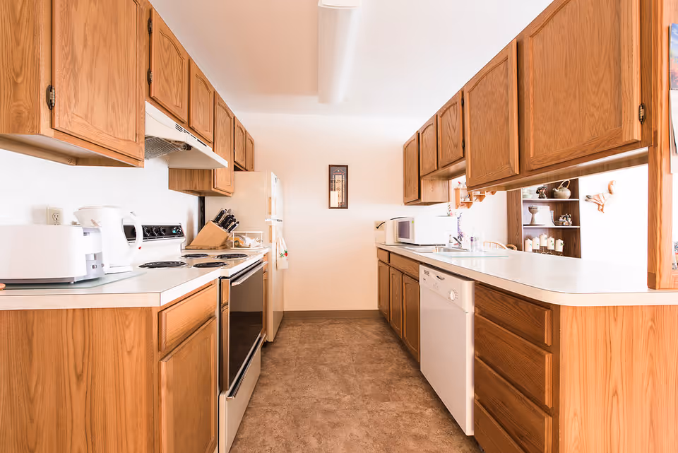 A bright kitchen with wooden cabinets on both sides, white countertops, and various appliances including a toaster, electric kettle, stove, refrigerator, microwave, and dishwasher. The floor is covered with a brown patterned linoleum, and there is a small framed picture on the far wall.