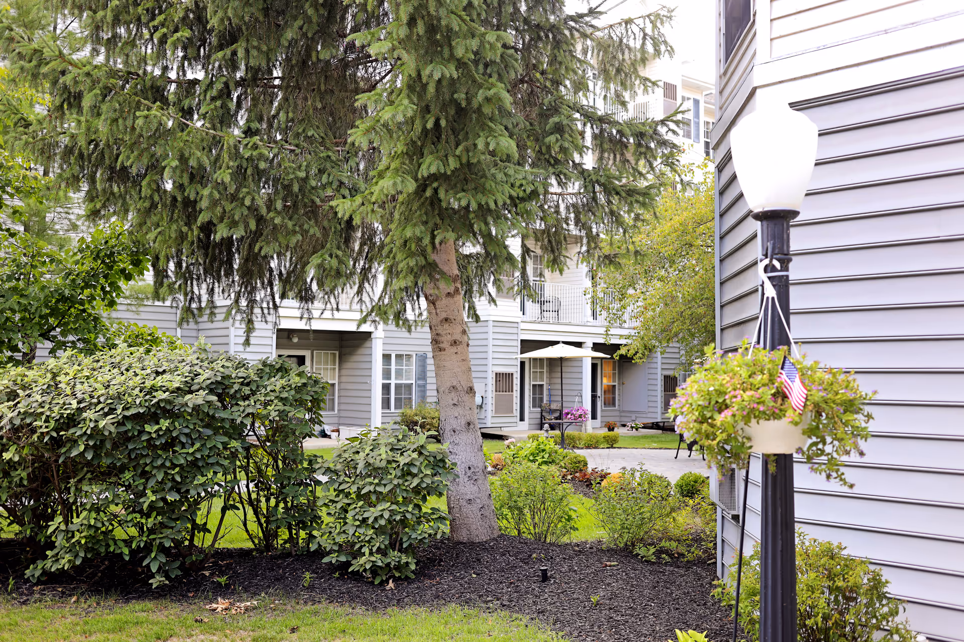 Outdoor garden area at Lockwood of Waterford with green bushes, a large tree, and a lamppost with a hanging flower pot. The background shows the exterior of a multi-story building with balconies and windows.