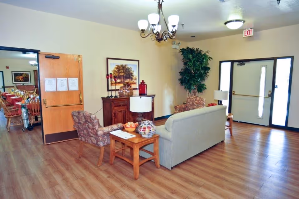 A cozy assisted living facility common area with a beige couch, two patterned armchairs, a wooden side table with a lamp and decorative items, a wooden cabinet with glass jars and candles, a framed painting on the wall, and a large potted plant near the entrance door. The floor is wood laminate, and there is a chandelier and ceiling light fixture. Through an open door, a dining area with tables and chairs is visible.
