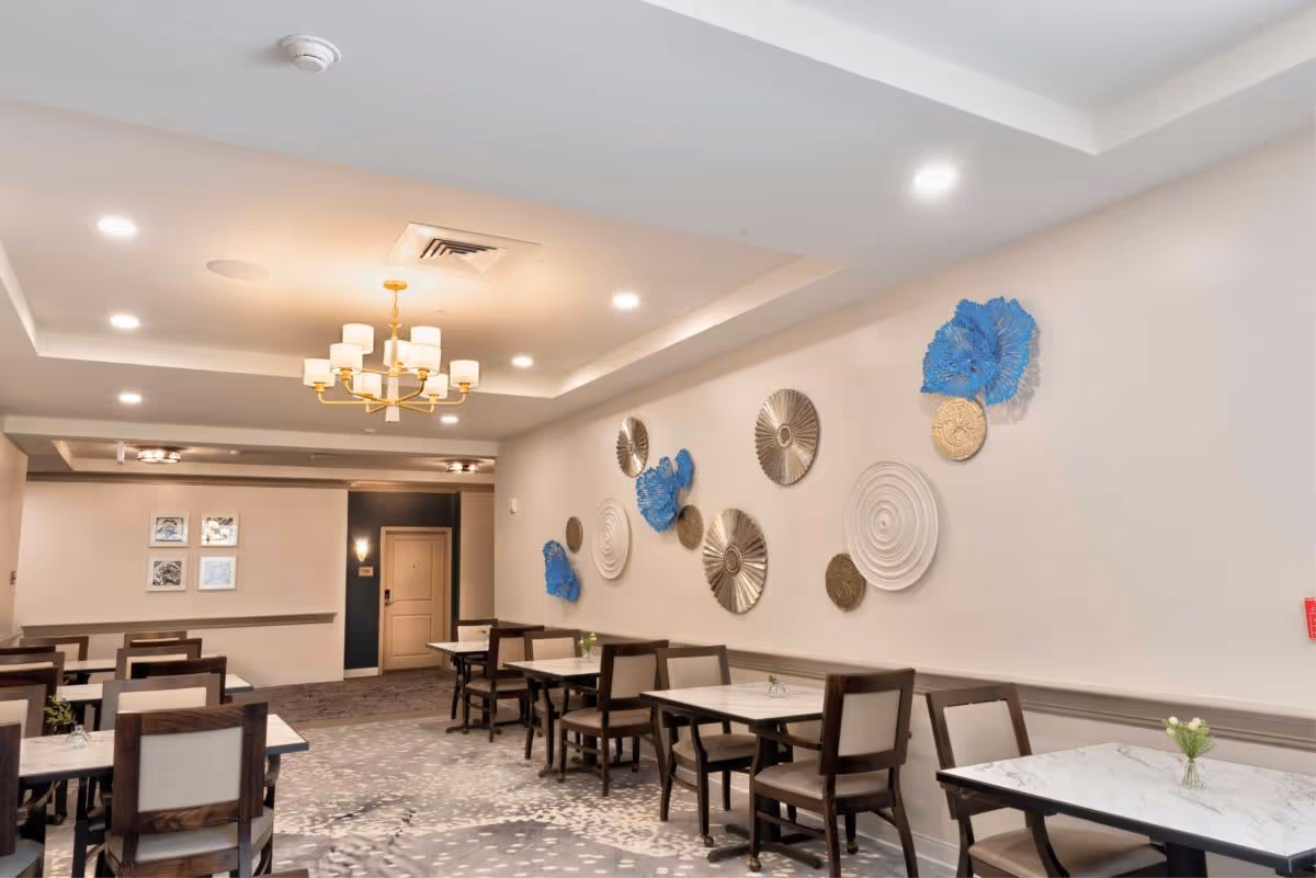 A dining room in Heartis Venice Assisted Living featuring several tables with chairs arranged neatly. The walls are decorated with circular art pieces in blue, white, and metallic colors. The ceiling has recessed lighting and a chandelier with multiple lampshades. The floor is carpeted with a patterned design.