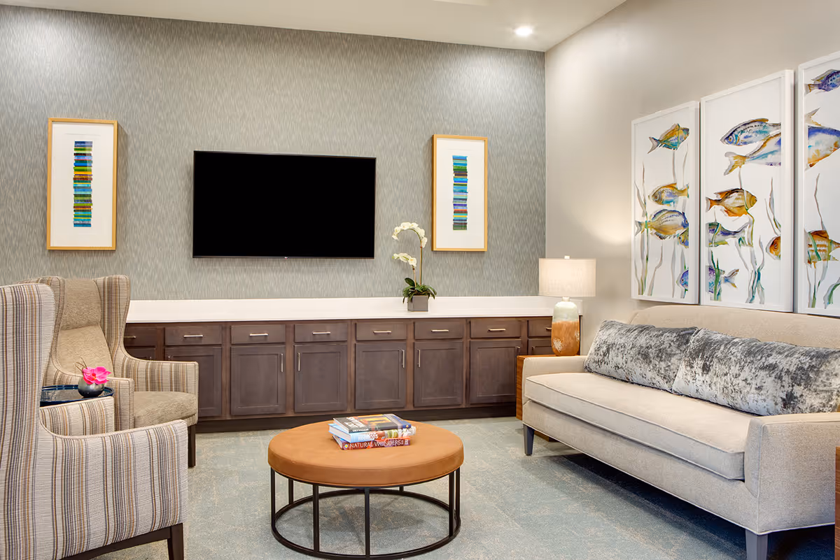 A cozy living room with a beige sofa adorned with two gray textured pillows, a round brown coffee table with books on top, and two striped armchairs. The wall behind the sofa features three framed paintings of colorful fish, while the opposite wall has a mounted flat-screen TV flanked by two framed abstract artworks. Below the TV is a long cabinet with dark wood doors and a white countertop, decorated with a small potted orchid and a table lamp.