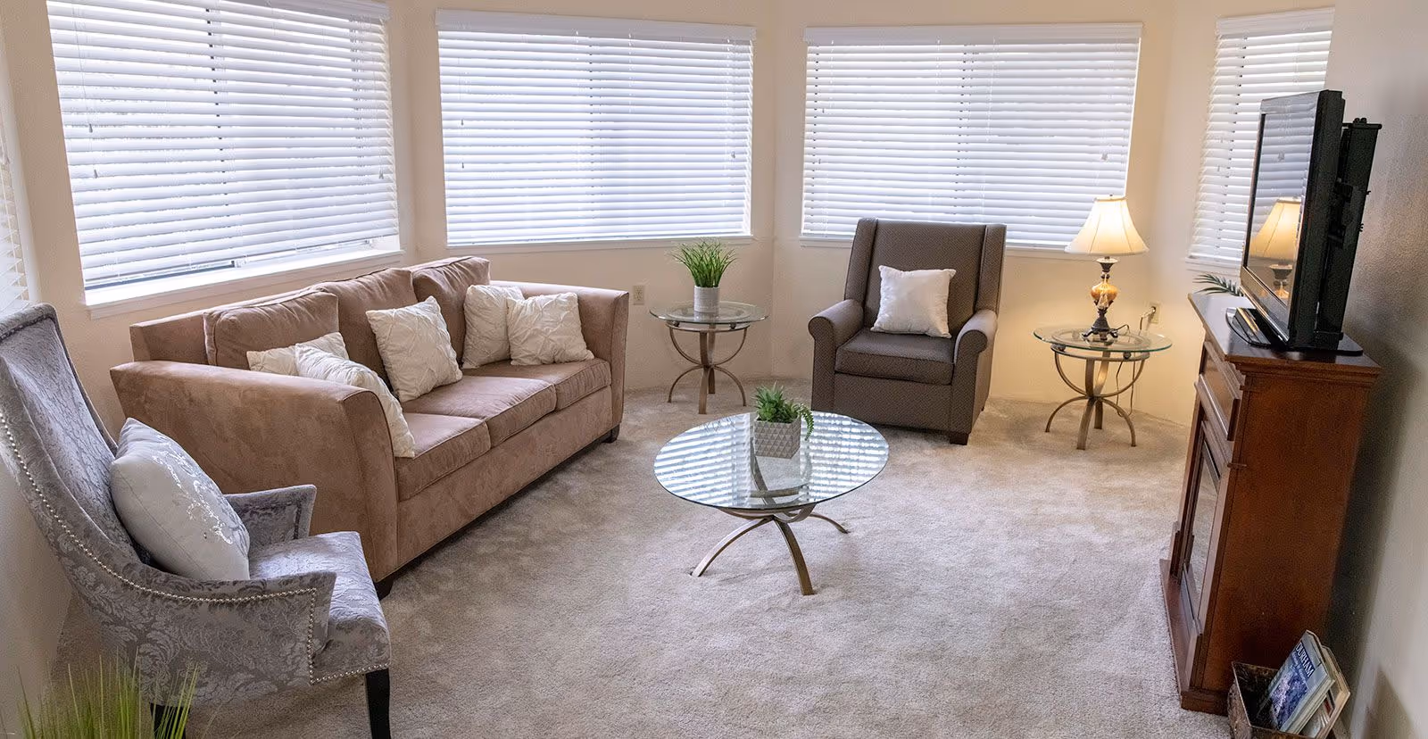 A bright living room with beige carpet, a brown sofa with white pillows, two armchairs with pillows, a glass coffee table with a small plant, two glass side tables with plants and a lamp, and a wooden TV stand with a flat-screen TV. The room has multiple windows with white blinds.