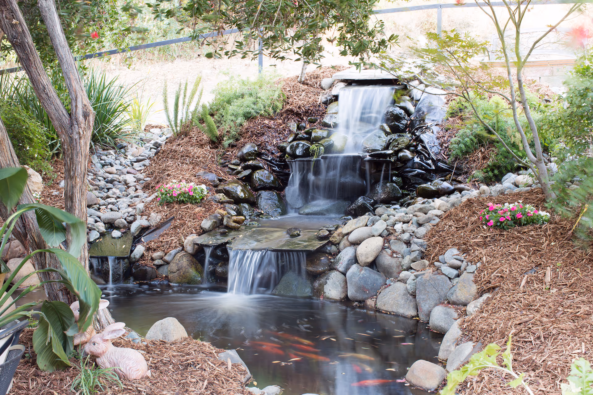 A serene outdoor garden area featuring a small multi-tiered waterfall flowing over rocks into a pond with visible fish. The surrounding landscape includes various plants, flowers, and mulch, with a decorative pink rabbit statue near the water's edge.