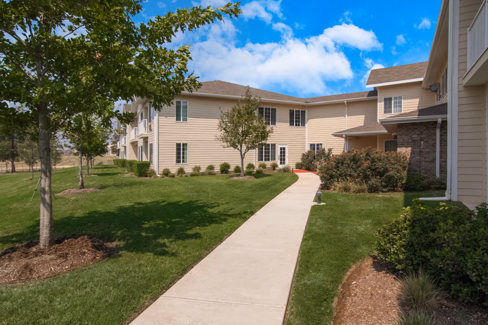 Pathway along beige senior living buildings with green lawns and trees under blue sky