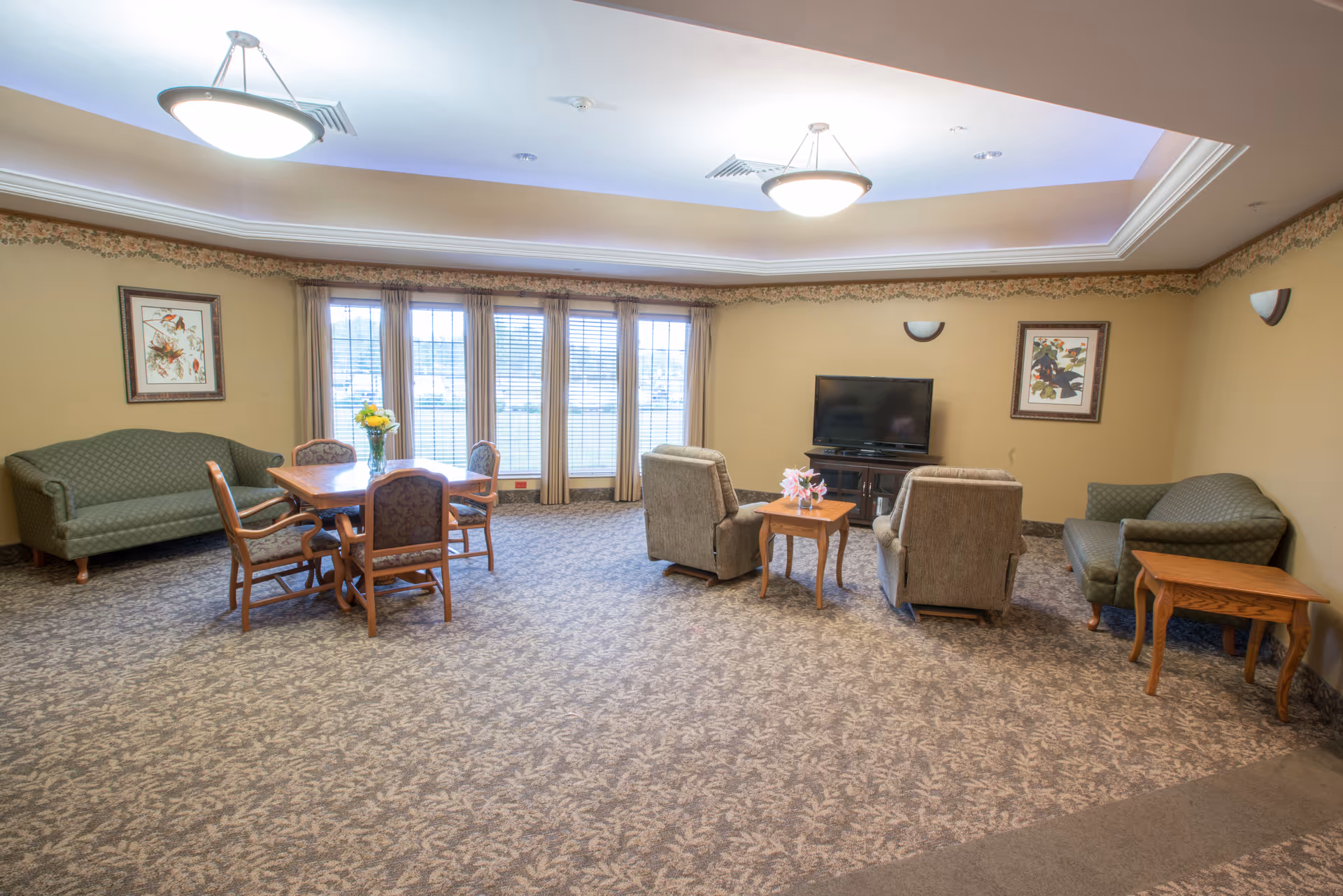A spacious common living area in a nursing and rehabilitation center featuring two green sofas, two armchairs facing a flat-screen TV on a wooden stand, a wooden table with four chairs and a vase of flowers, beige walls with framed artwork, large windows with curtains, and patterned carpet flooring.