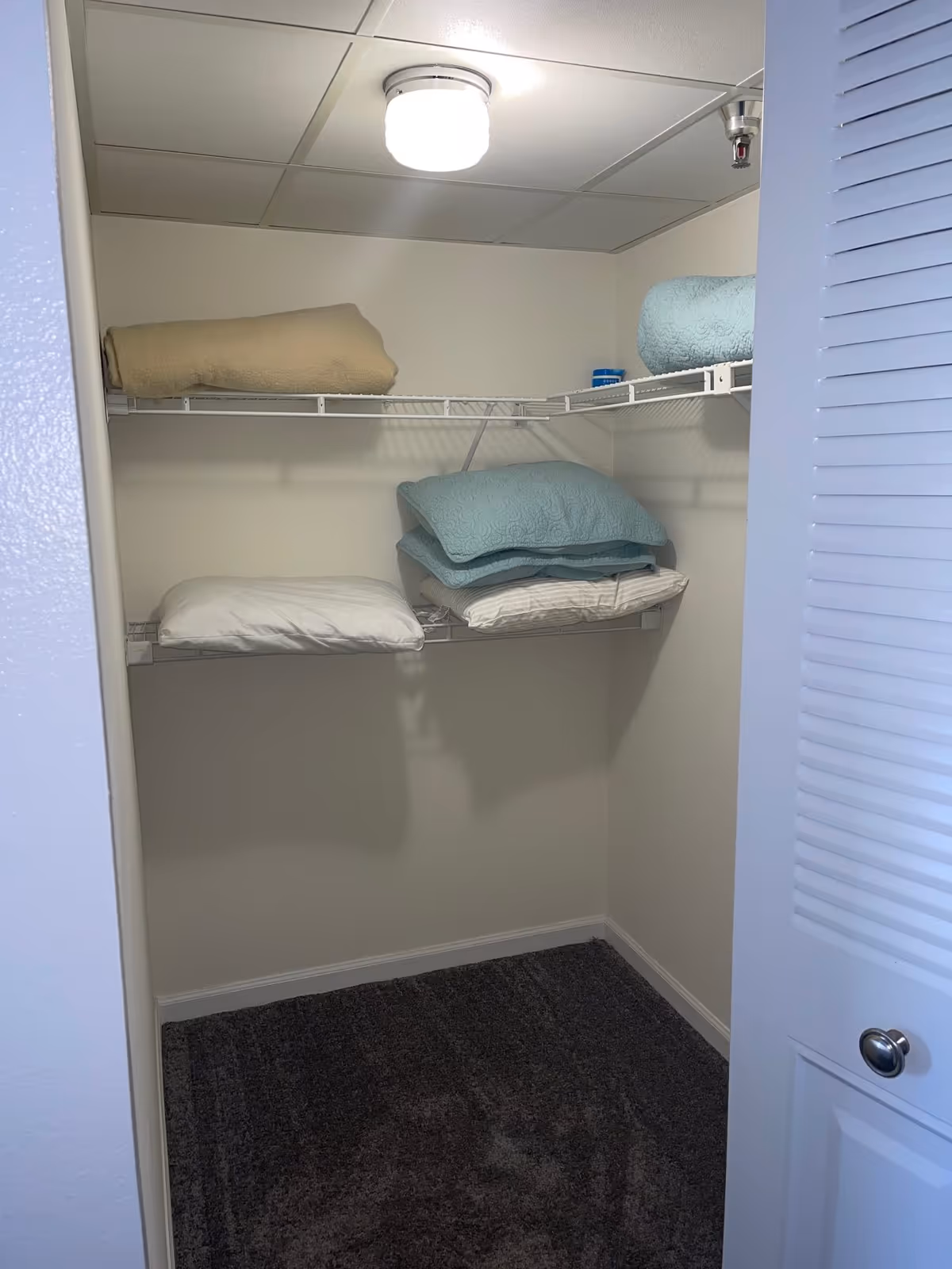 A small closet with white wire shelves holding folded blankets and pillows. The closet has beige walls, a dark carpeted floor, a white ceiling light, and a partially open white louvered door.