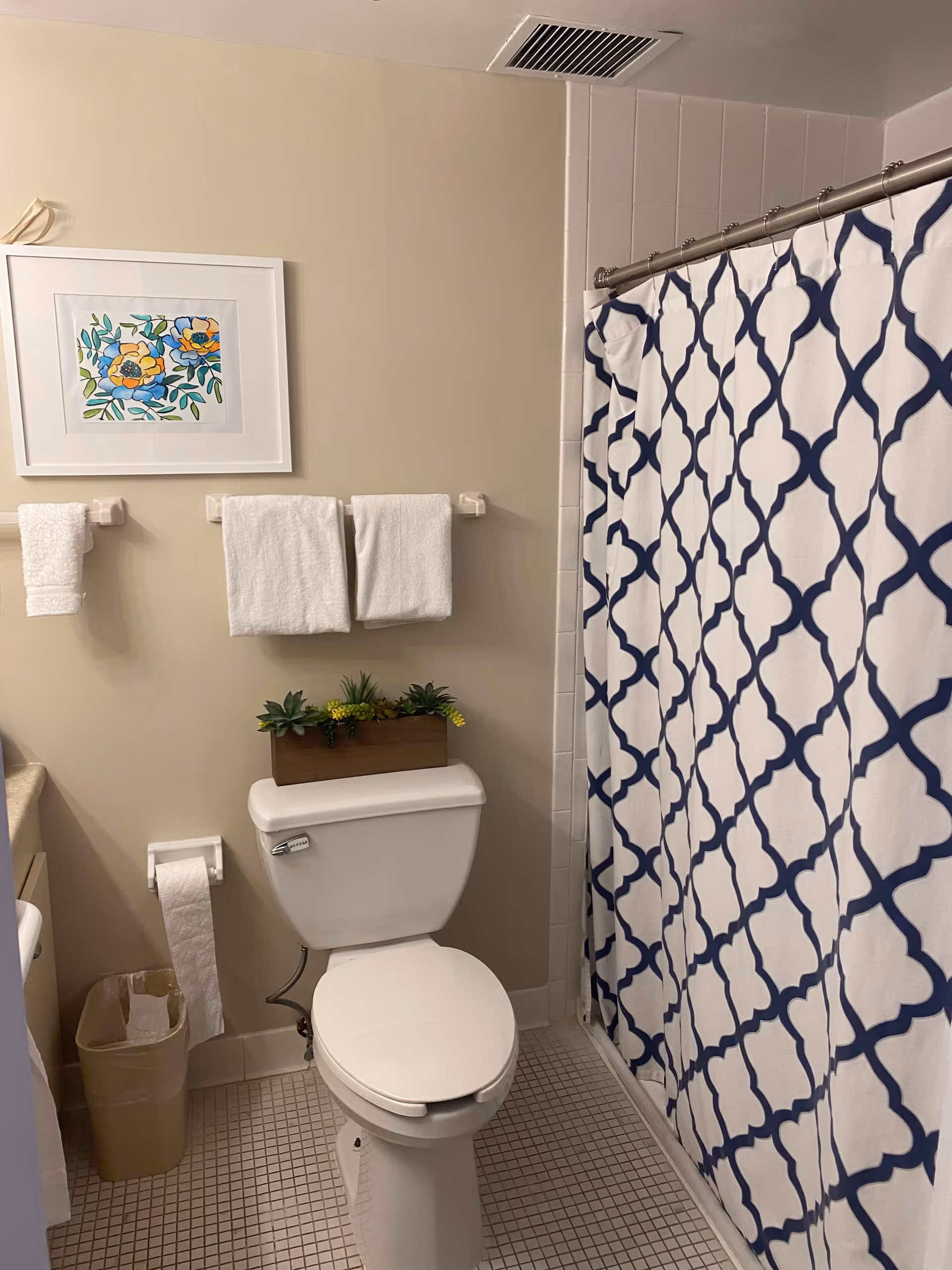 A bathroom with a white toilet, a small rectangular planter with green and yellow artificial plants on top of the toilet tank, a white and blue patterned shower curtain, three white towels hanging on the wall, a small trash can, and a framed floral artwork on the wall.
