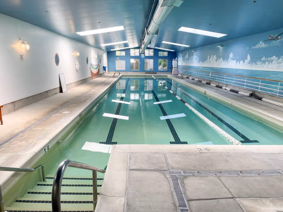 Indoor swimming pool with clear water, lane dividers, and steps leading into the pool. The walls are decorated with a mural of a sky and ocean scene, and there are windows at the far end letting in natural light. The ceiling is painted blue with exposed ductwork and overhead lights.