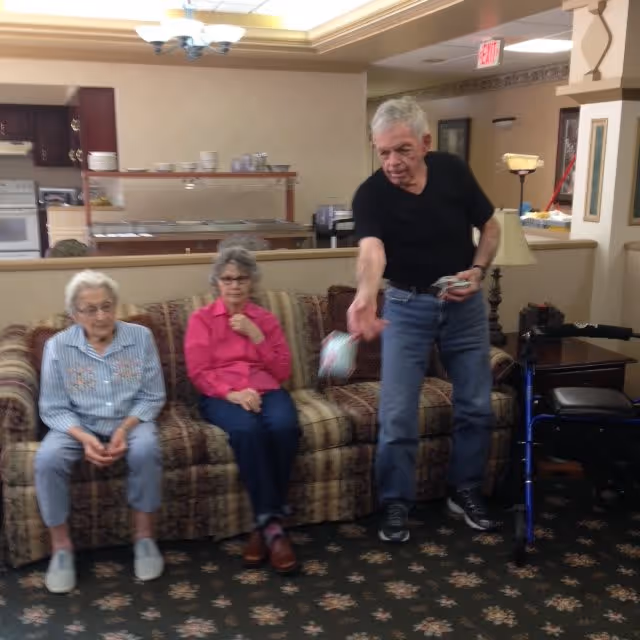 Three elderly individuals in a living room area of a senior living facility. Two women are seated on a patterned sofa, one wearing a striped shirt and the other in a bright pink shirt. A man in a black shirt and jeans is standing and appears to be tossing or handing something. The room has a patterned carpet, a walker nearby, and a kitchen area visible in the background.