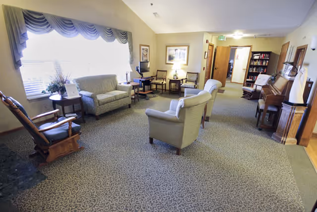 A well-lit common area in a senior living facility with multiple upholstered chairs, a loveseat, a wooden rocking chair, side tables with lamps, framed artwork on the walls, a bookshelf, and a piano. The room has carpeted flooring and large windows with decorative valances allowing natural light to enter.