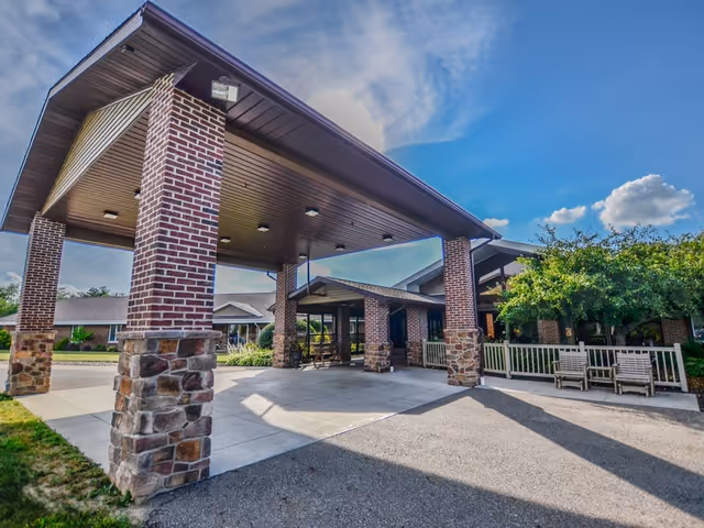 Entrance of a single-story brick building with a covered driveway supported by brick and stone pillars, outdoor benches, and a clear blue sky with some clouds.