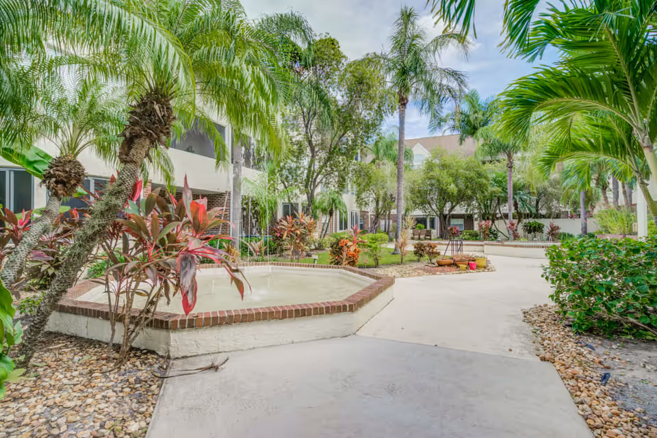 Landscaped courtyard with palm trees, tropical plants, a brick-edged fountain and paved walkways in front of the building.