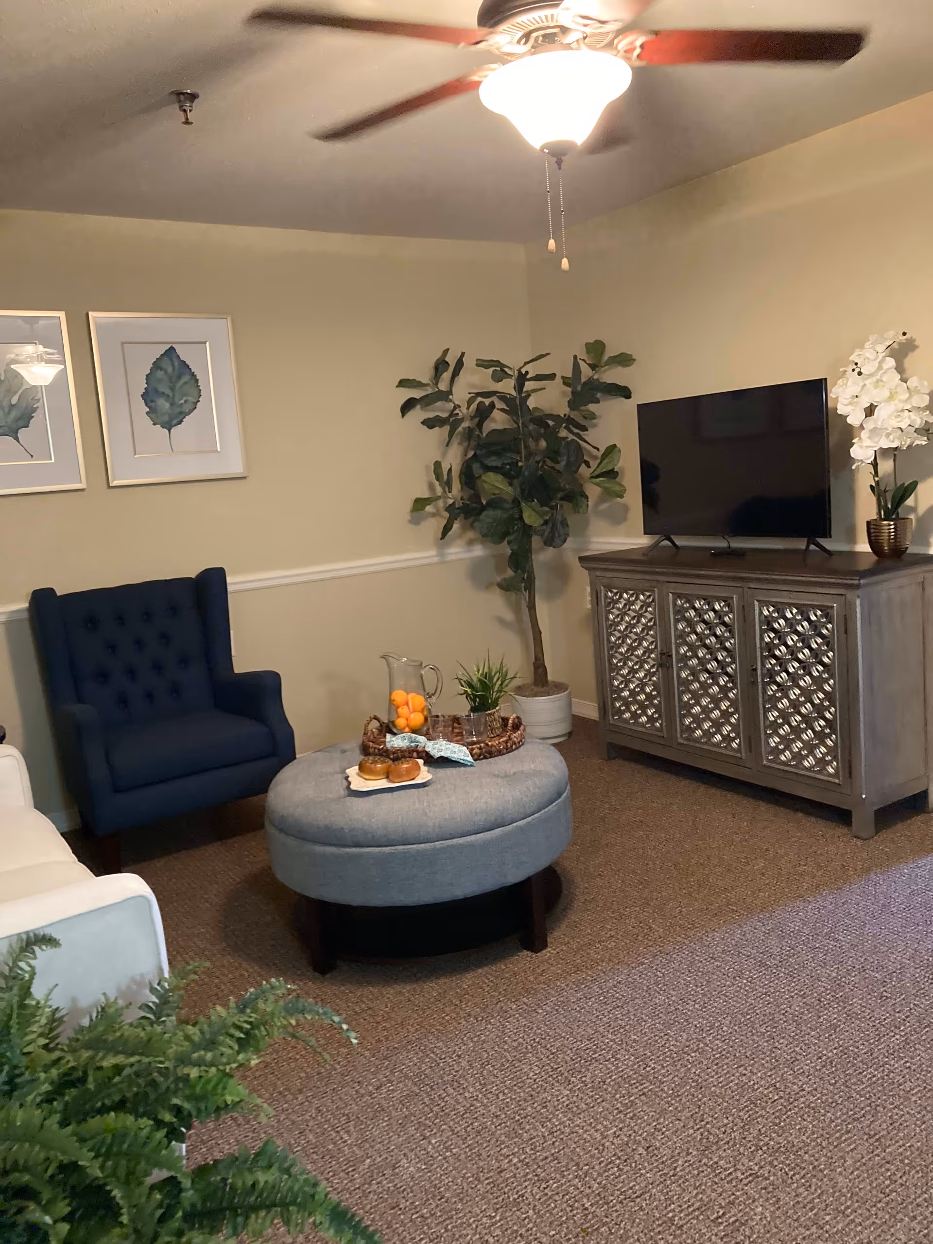 A cozy living room with a blue armchair, a round gray ottoman with a tray holding a pitcher of orange drink, two glasses, and two bagels. There is a TV on a decorative wooden cabinet, a tall green plant in a white pot, and white flowers in a gold vase. Two framed leaf prints hang on the beige wall, and a ceiling fan with a light is on the ceiling.