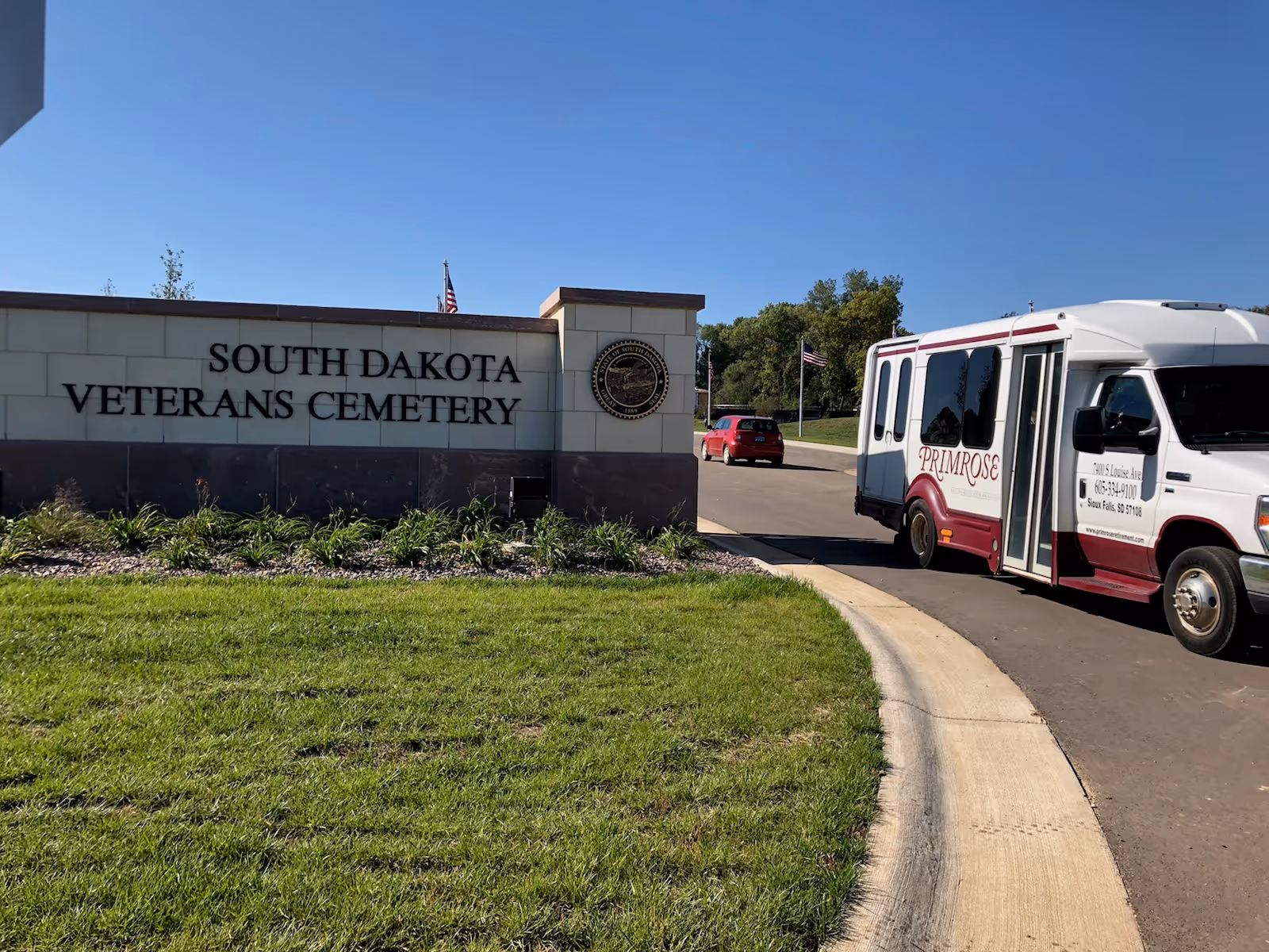 Entrance sign for South Dakota Veterans Cemetery with a white and maroon Primrose senior living shuttle bus driving on the road beside it under a clear blue sky.