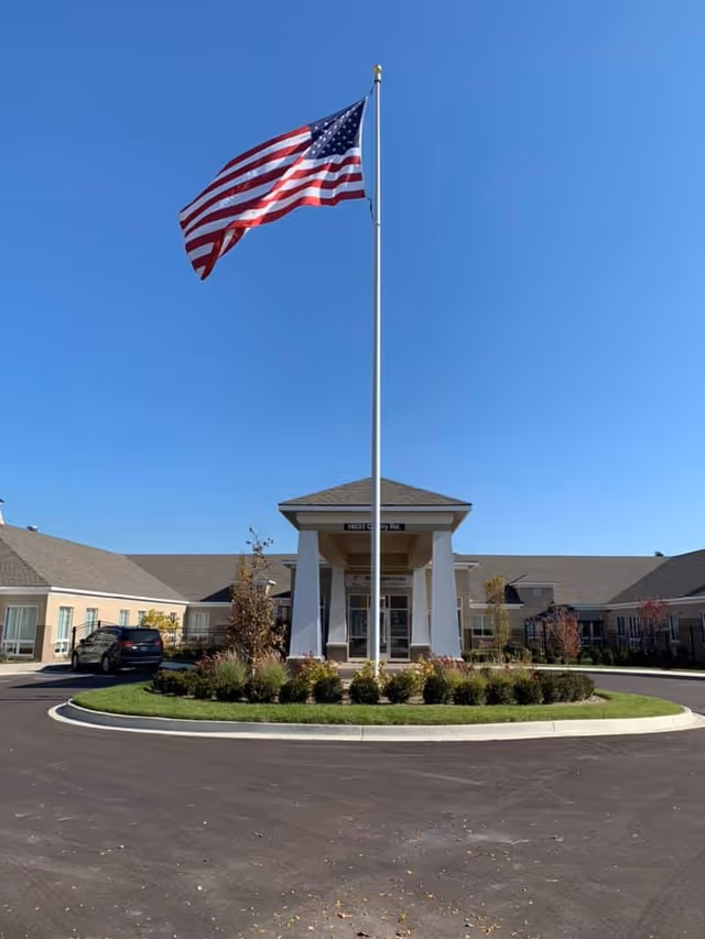 Front entrance of a senior living building with a tall flagpole flying an American flag over a circular driveway.