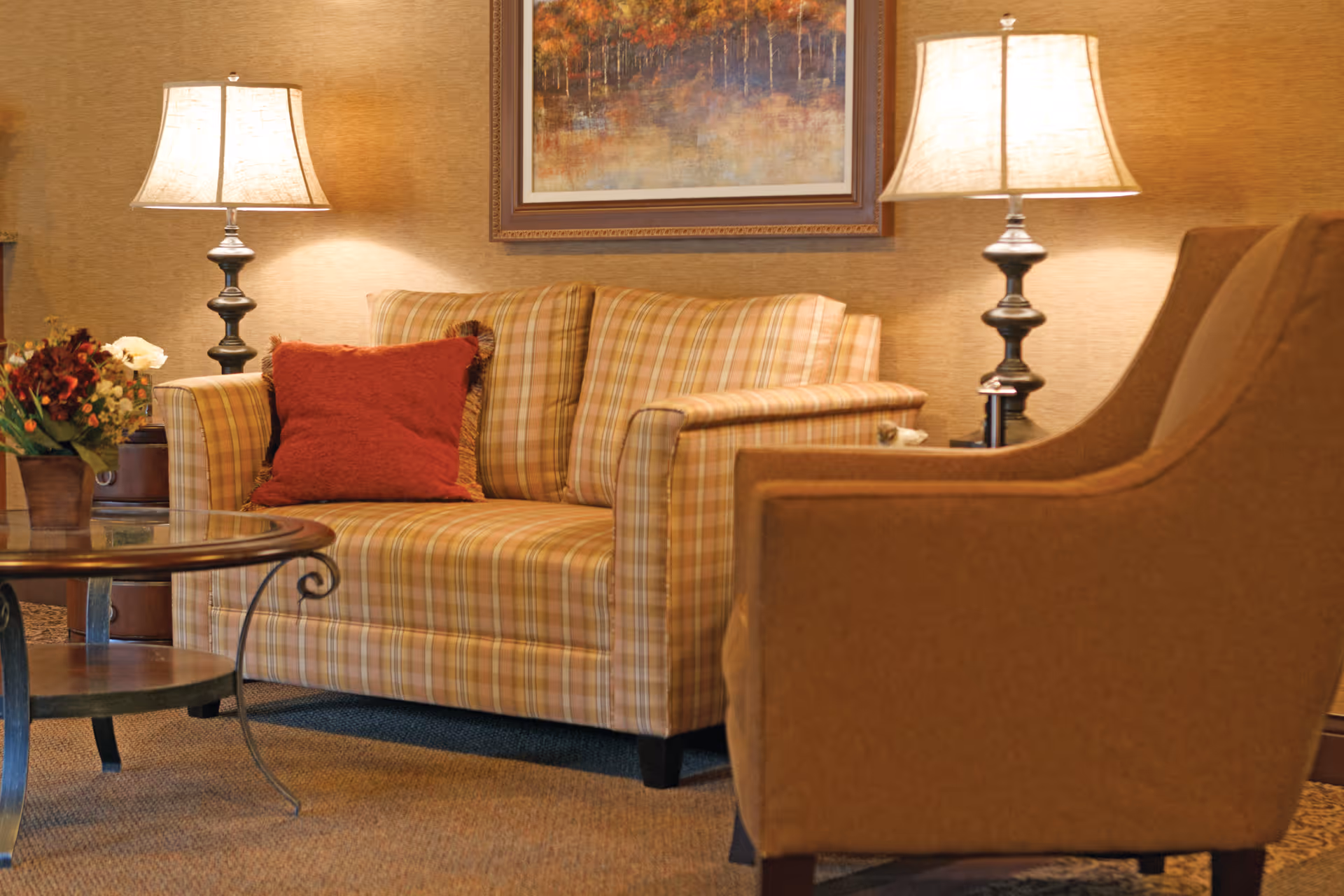 Cozy lounge area with a plaid loveseat, brown armchair, two table lamps, coffee table and wall art.