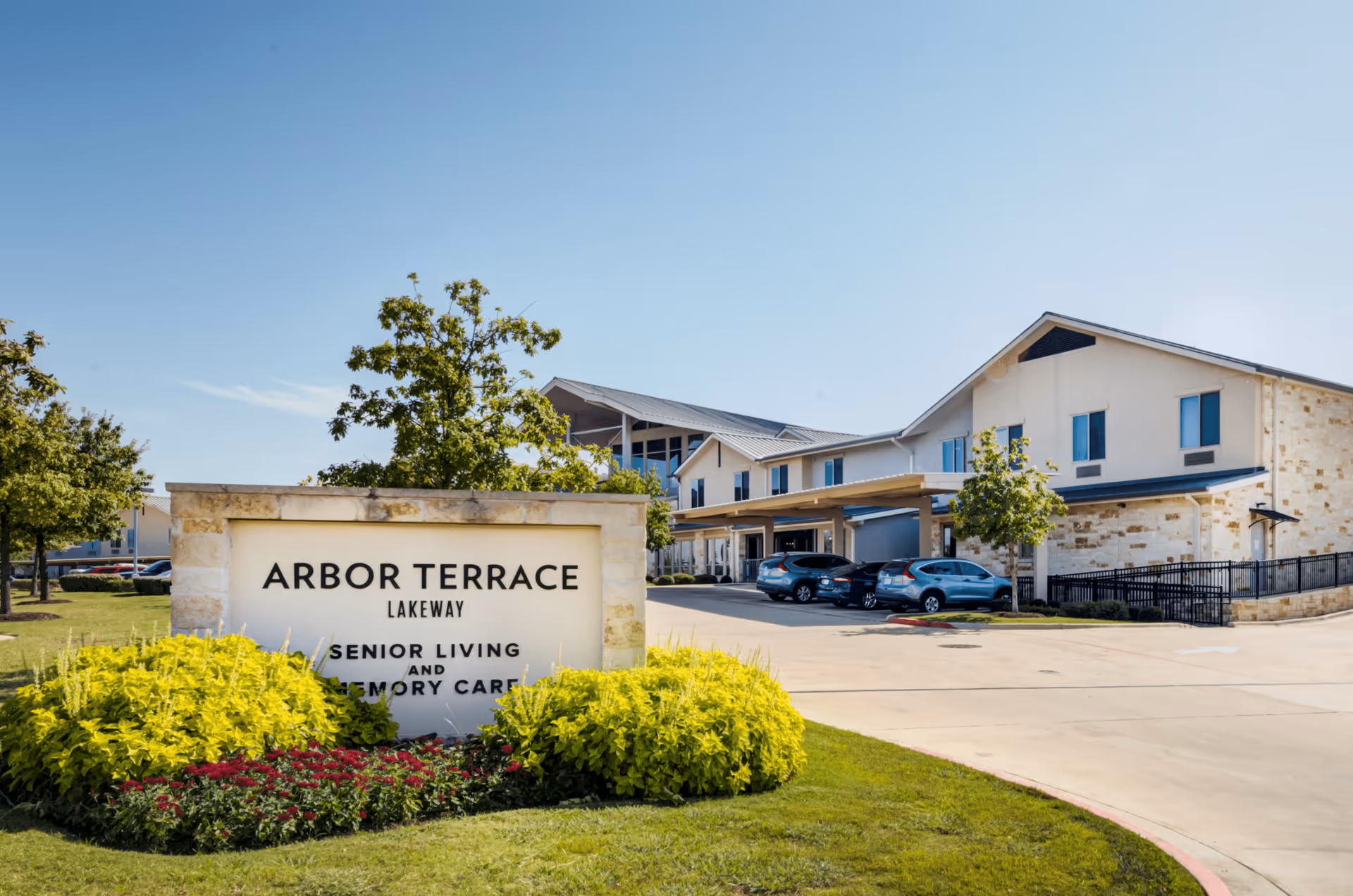 Exterior view of Arbor Terrace Lakeway senior living and memory care facility with a stone sign in the foreground surrounded by green shrubs and flowers, and a two-story building with parked cars under a clear blue sky.