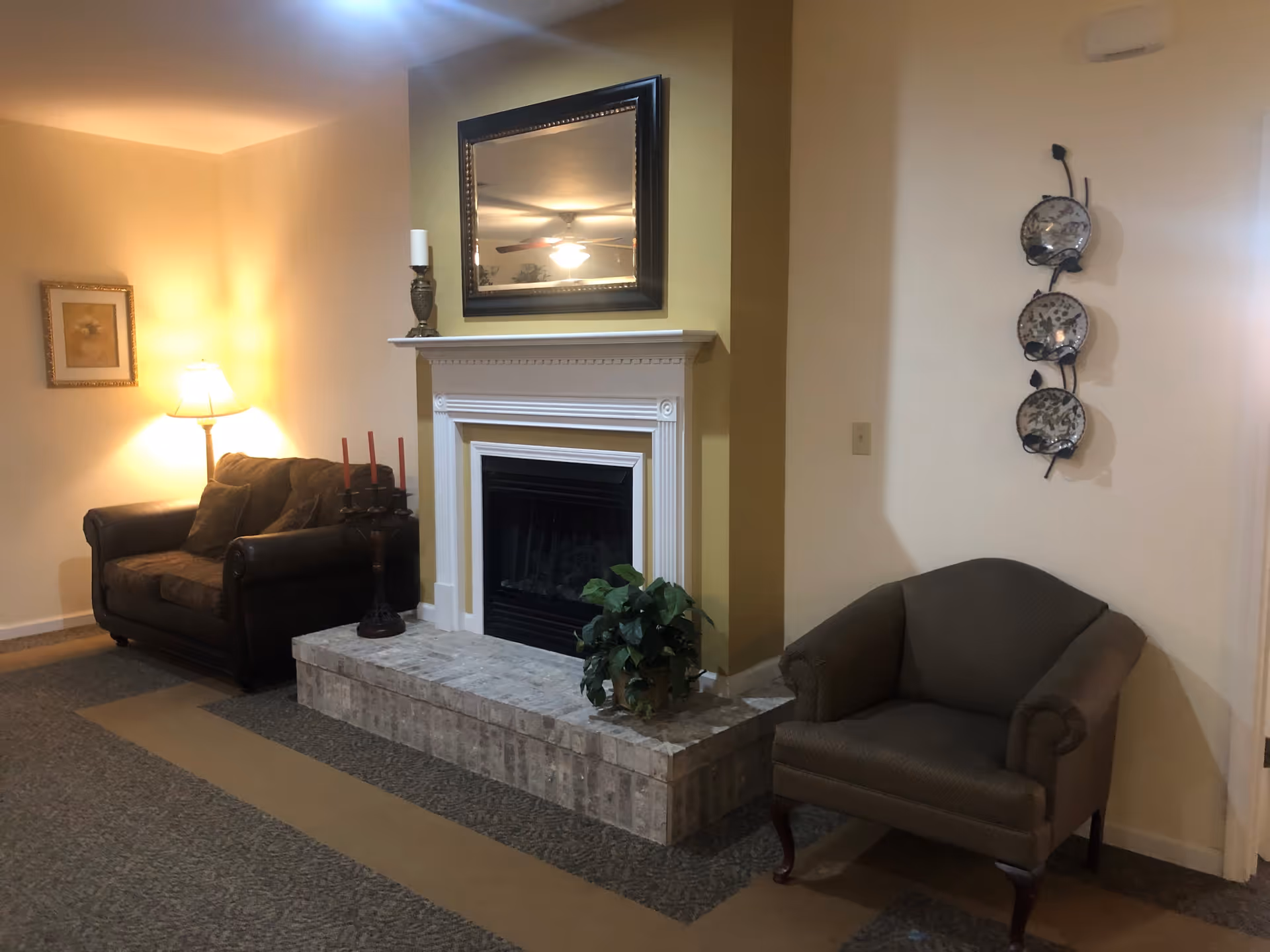 Living-room seating area with a fireplace and mantel, a mirror above, an armchair and loveseat, lamp, and wall decor.