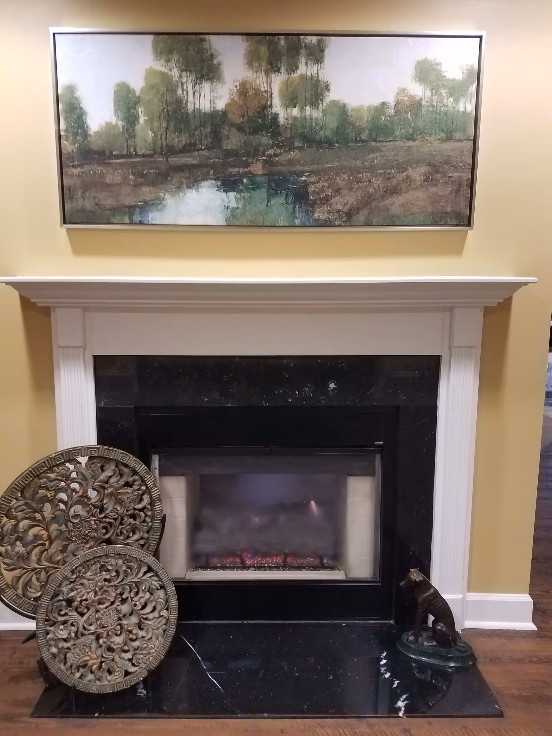 A cozy indoor fireplace with a white mantel and black marble surround. Above the fireplace hangs a large framed painting depicting a serene landscape with trees and a pond. On the hearth, there are two decorative round metal plaques with intricate designs leaning against the left side, and a small bronze dog statue sitting on a green base on the right side.