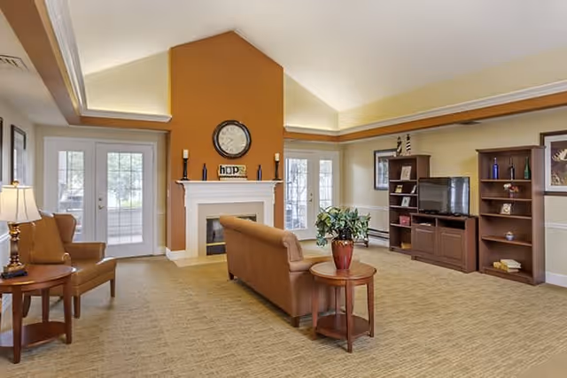A spacious living room with beige carpet and walls painted in warm tones. The room features a brown sofa facing a fireplace with a clock and decorative items on the mantel. There are two armchairs with a side table and lamp on the left side. On the right side, there is a wooden entertainment center with a TV and bookshelves. French doors with glass panes lead outside, allowing natural light to fill the room.