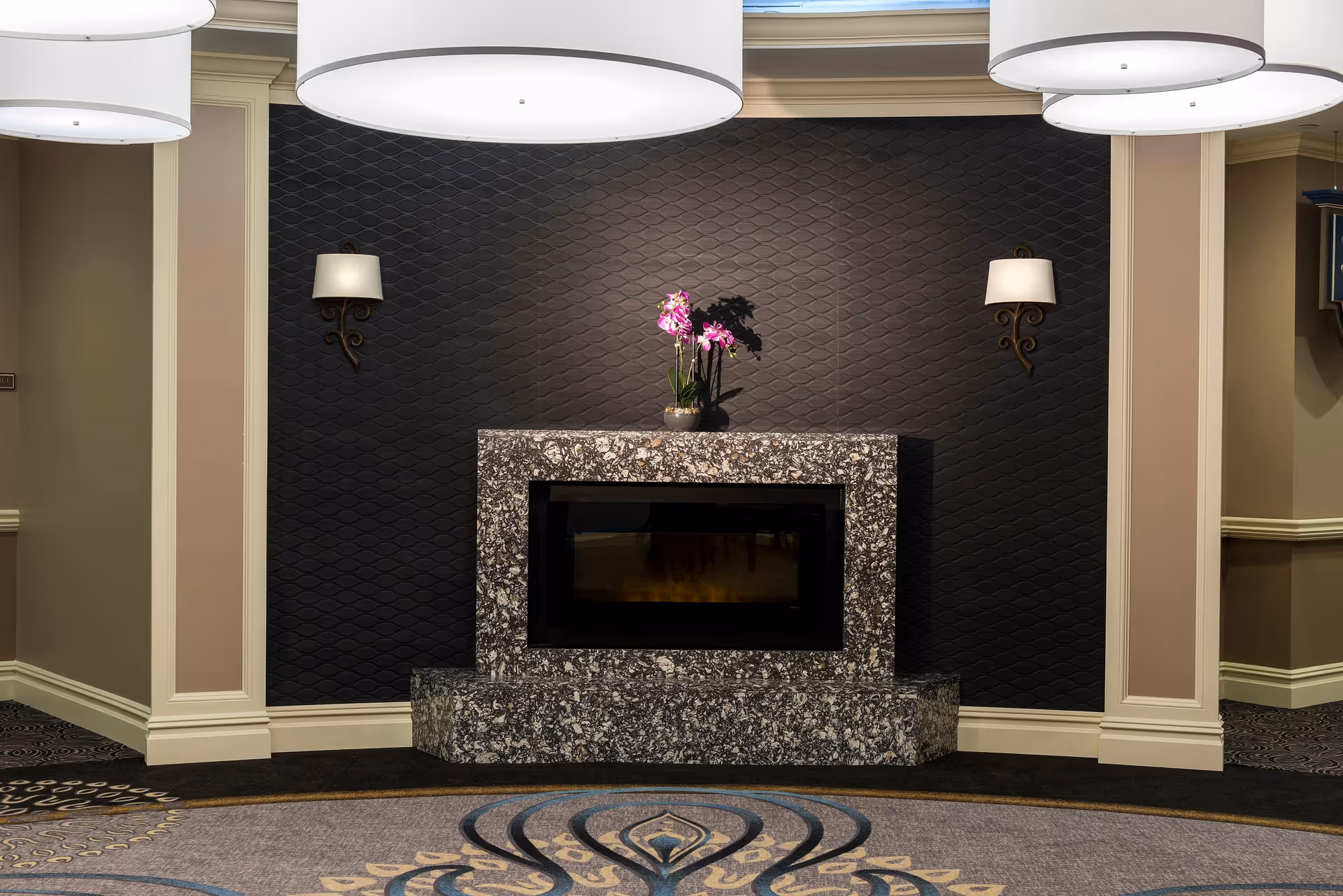 A modern indoor fireplace with a speckled stone surround set against a textured dark wall. On top of the fireplace is a small pot with pink orchids. Two wall-mounted lamps with white shades flank the fireplace. The room features beige walls with white trim and a patterned carpet with blue and beige designs. Large round ceiling lights hang above.