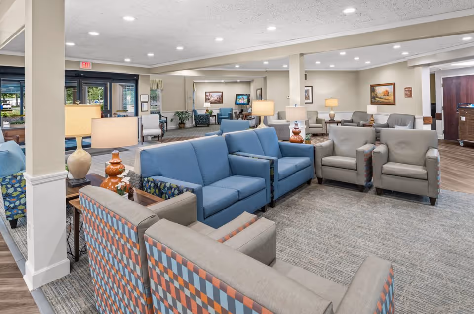 A spacious and well-lit living room area in a senior living facility with multiple seating arrangements including blue and patterned armchairs and sofas. The room features several table lamps, framed artwork on the walls, and large windows near the entrance allowing natural light to enter.