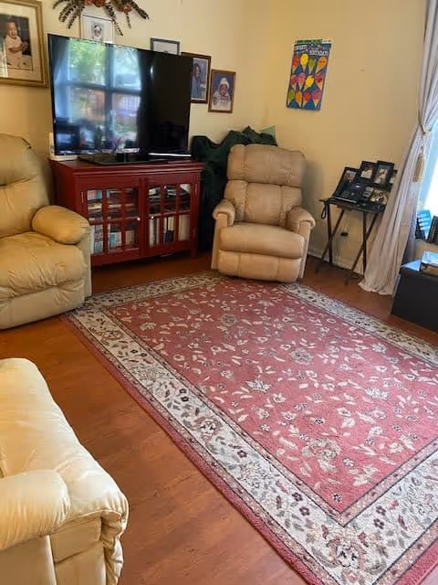 A cozy living room with beige recliner chairs, a large flat-screen TV on a red wooden cabinet, framed pictures on the walls, a patterned red area rug on wooden flooring, and a small side table with photo frames near a window with white curtains.
