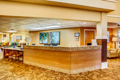 Reception area in a senior living facility with a wooden front desk, two colorful paintings on the wall behind the desk, and a seating area with chairs and a table to the left. The space has warm lighting and carpeted floors with a patterned design.