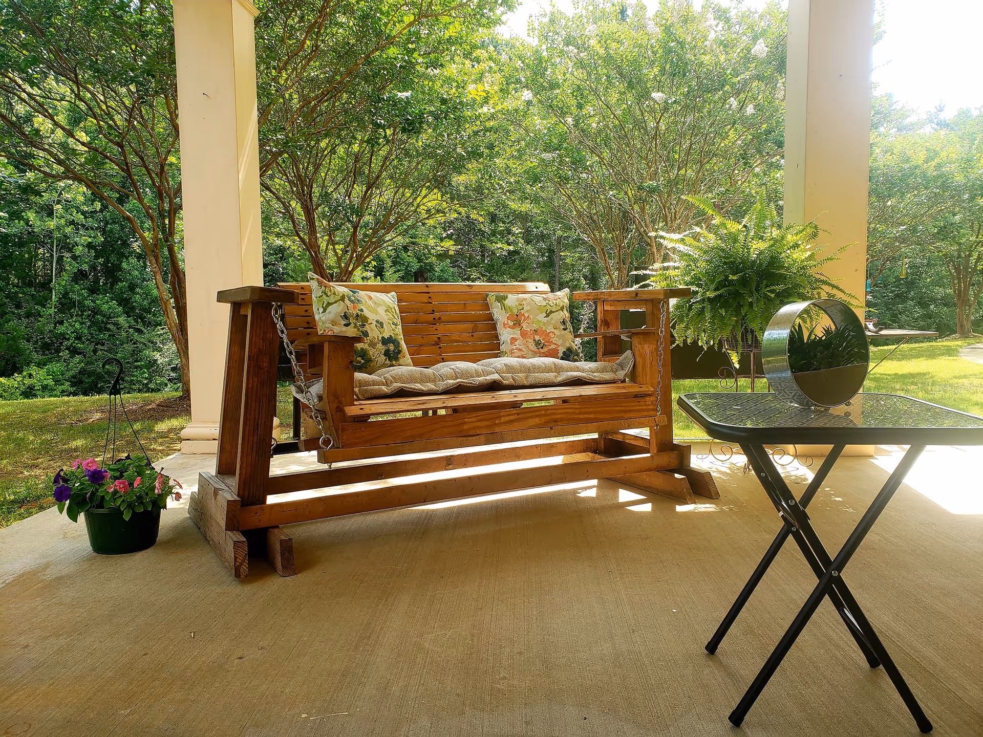 A wooden porch swing with floral cushions and a padded seat cushion sits on a covered patio. Next to it is a small glass-top table with a decorative circular mirror and a potted fern plant. In the background, there are green trees and a grassy area under bright sunlight.