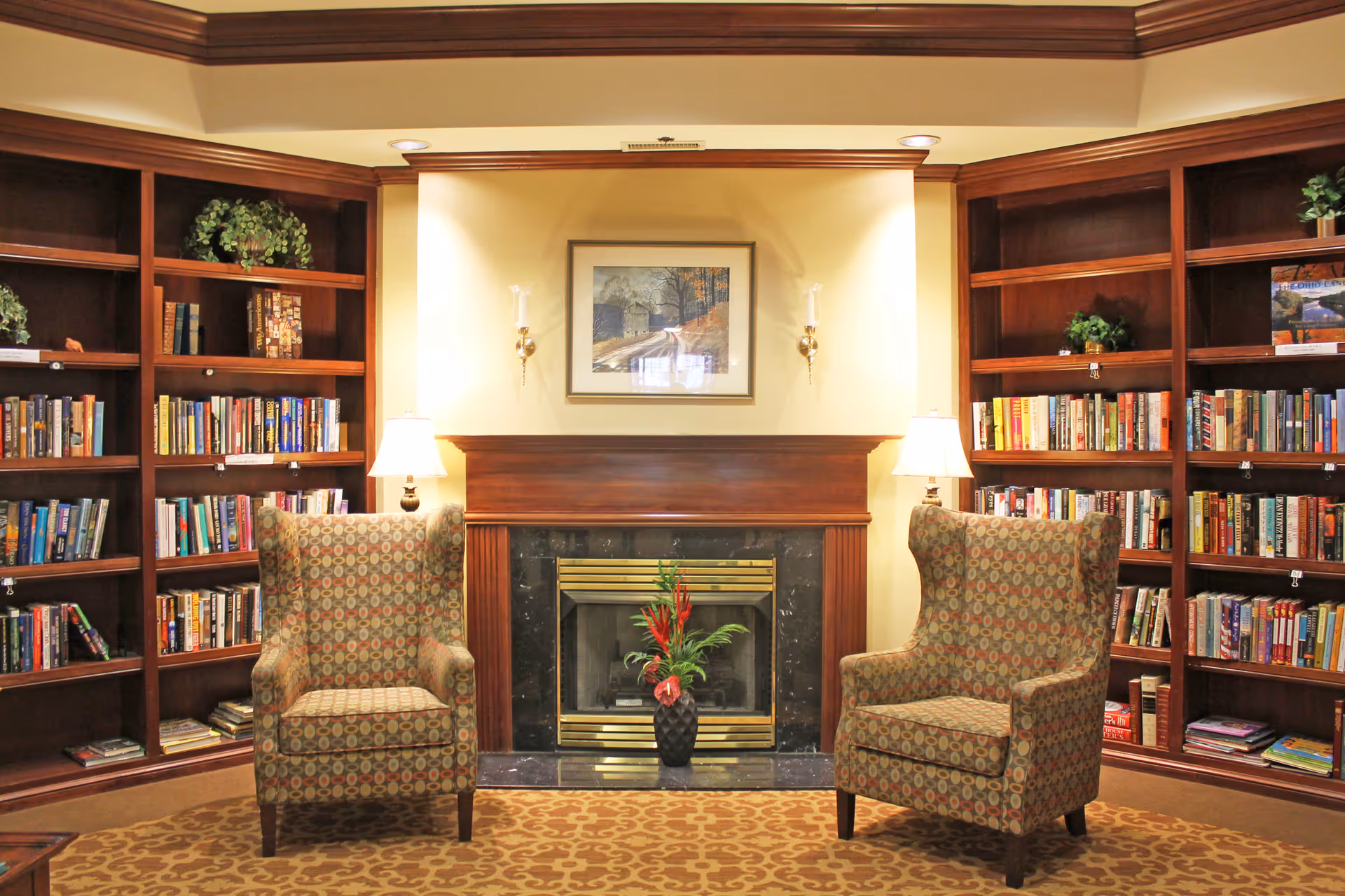 A cozy reading area with two patterned armchairs facing a fireplace. The fireplace has a dark marble surround and a wooden mantel with a framed picture above it. On either side of the fireplace are built-in wooden bookshelves filled with books and some decorative plants. Two table lamps are positioned behind the armchairs, providing warm lighting.
