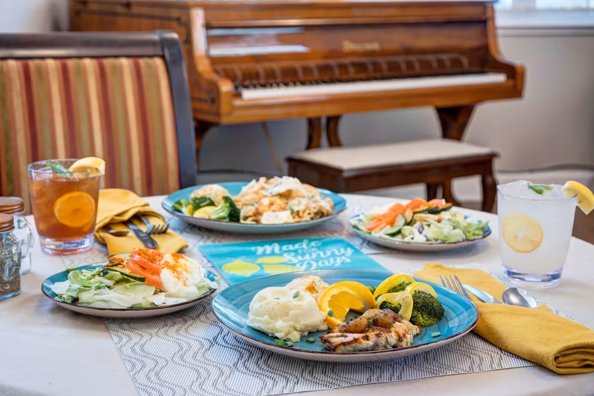 A dining table set with plates of food including grilled chicken with broccoli and mashed potatoes, a salad with tomatoes and lettuce, and another plate with vegetables. There are two drinks, one iced tea with lemon and one lemonade with a lemon slice and mint. The table has yellow napkins and silverware, and a piano is visible in the background.