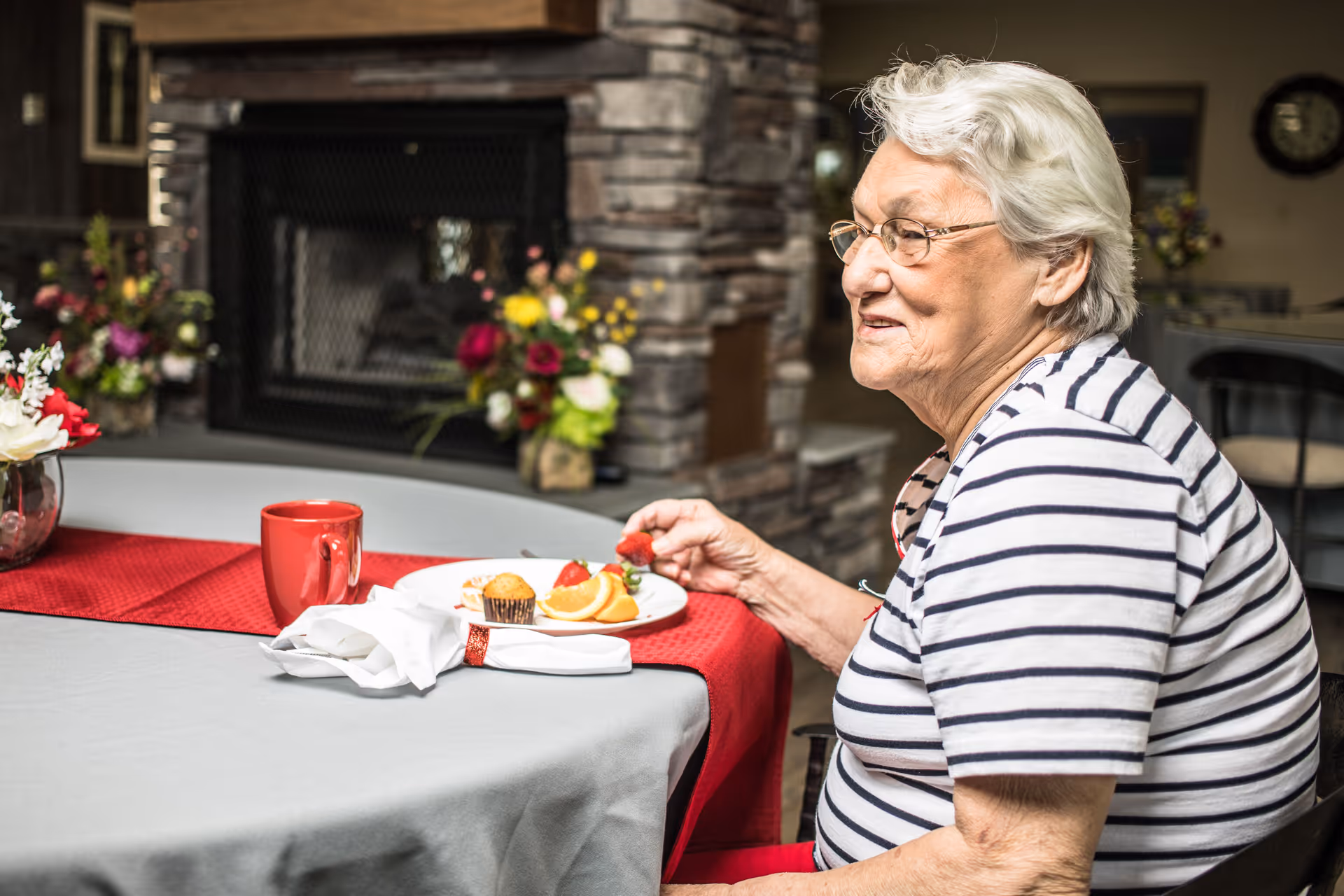 An elderly woman with white hair and glasses, wearing a striped shirt, sitting at a round table with a gray tablecloth and red runner, holding a strawberry. On the table are a plate with a muffin, orange slices, and strawberries, a red mug, and a folded napkin. In the background is a stone fireplace with flower arrangements on either side.