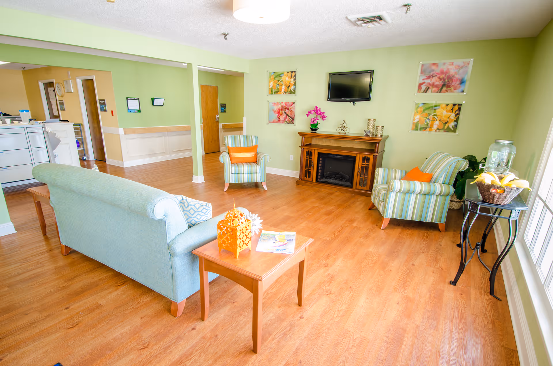 A bright and cozy living room with light green walls and wooden flooring. The room features a light blue sofa, two striped armchairs with orange cushions, a wooden coffee table with decorative items, a wooden electric fireplace, a wall-mounted TV, and colorful floral artwork on the walls. A small table near the window holds a basket of bananas and a water dispenser.