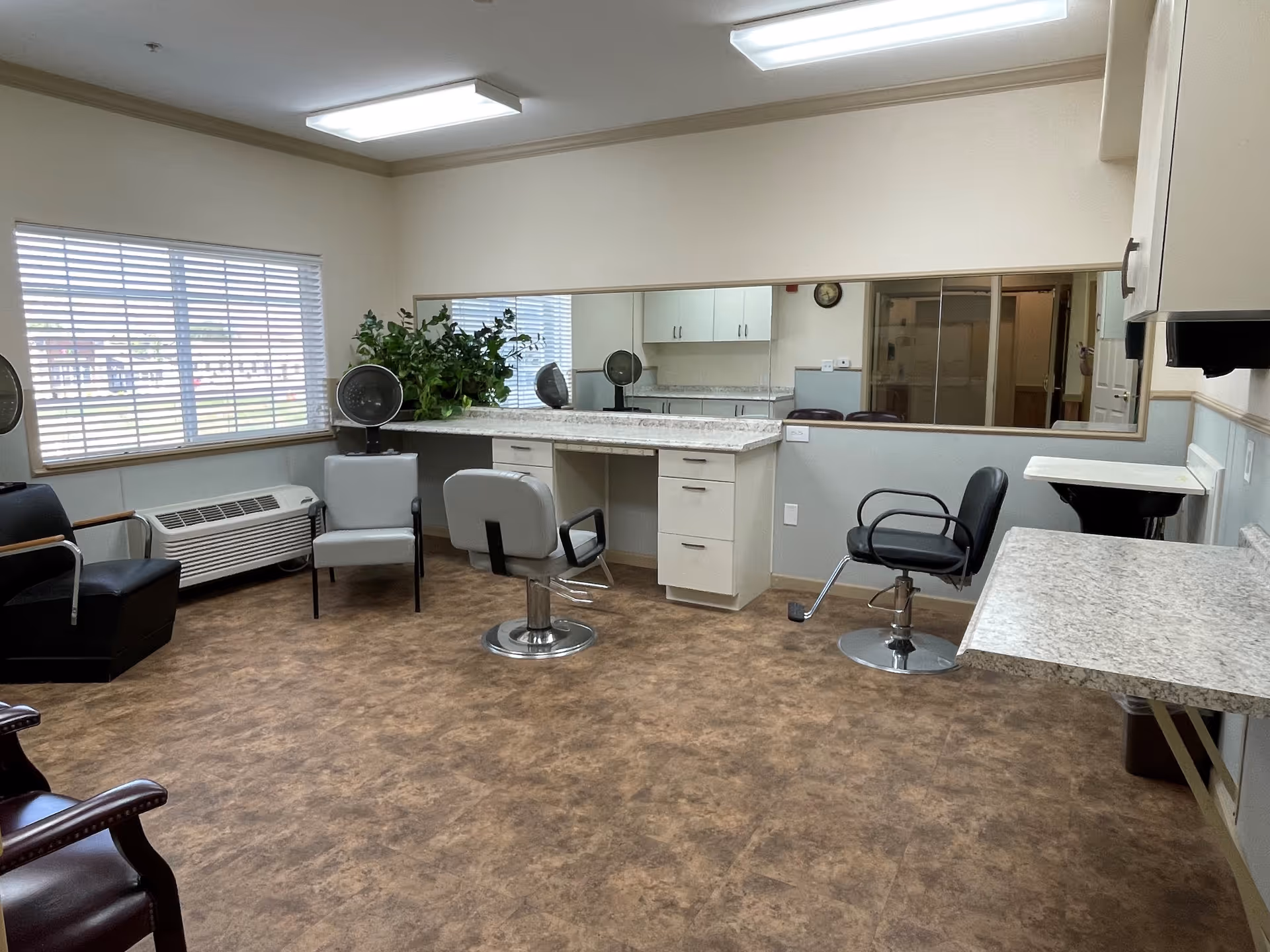 Interior view of a hair salon area in an assisted living facility with several salon chairs, a large mirror on the wall, a window with blinds, a plant, and a countertop with drawers.