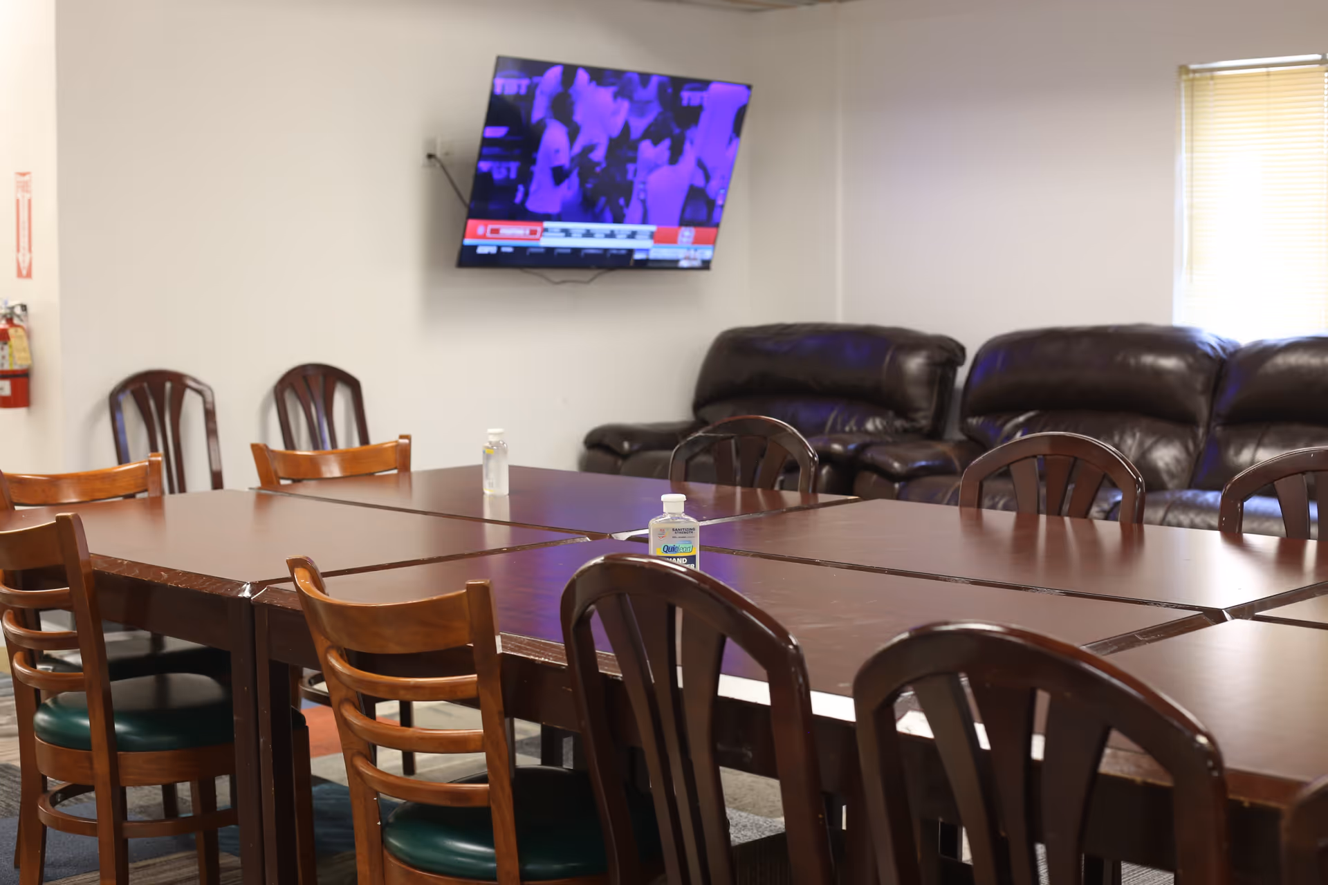 A room with a large rectangular wooden table surrounded by wooden chairs with green cushions. Two bottles of hand sanitizer are placed on the table. In the background, there are dark brown leather sofas and a wall-mounted TV displaying a sports event. A window with closed blinds is on the right side of the room.