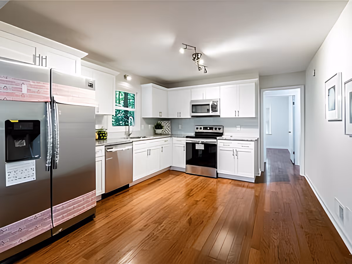 A modern kitchen with white cabinets, stainless steel appliances including a refrigerator, dishwasher, stove, and microwave. The kitchen has wooden flooring and a window above the sink letting in natural light. There are two framed pictures on the right wall and a hallway leading to another room.