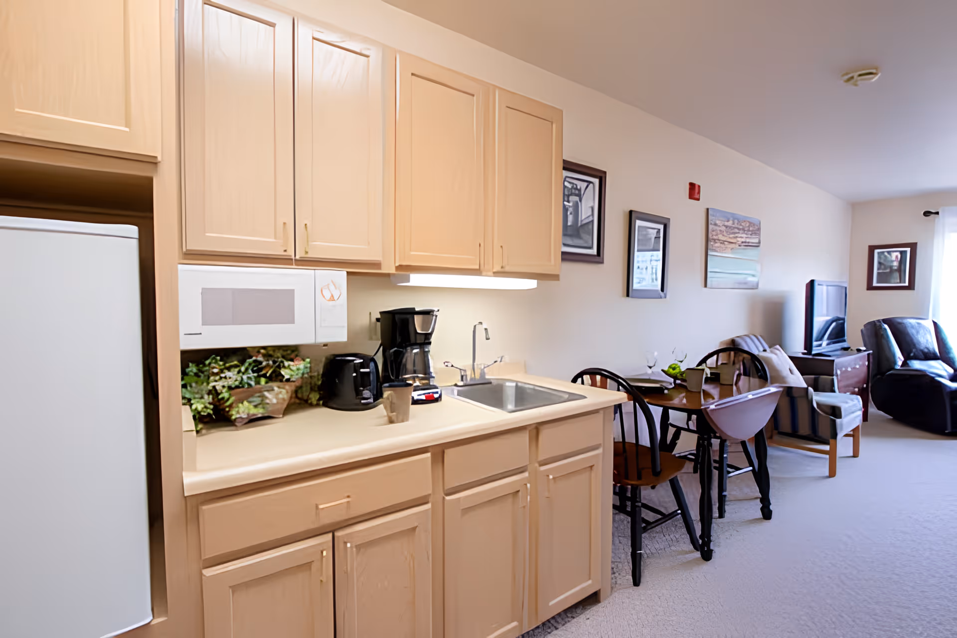 Interior view of a senior living facility apartment showing a small kitchen area with light wood cabinets, a microwave, coffee maker, toaster, and sink. Adjacent to the kitchen is a dining table with chairs, and further back is a living area with armchairs, a TV, and framed artwork on the walls.
