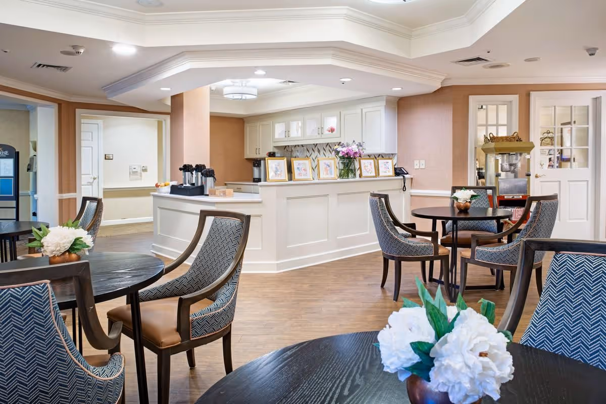 A bright and welcoming dining area in a senior living facility with round dark wood tables and patterned upholstered chairs. White flowers in small vases decorate the tables. In the background, there is a white counter with framed artwork and a vase of flowers, along with coffee dispensers and a popcorn machine near a door with glass panels.