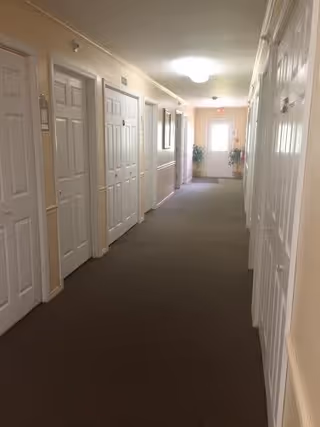 A long indoor hallway with beige walls and brown carpet. Multiple white doors line both sides of the hallway. At the end of the hallway, there is a door with a window letting in natural light and some potted plants nearby.