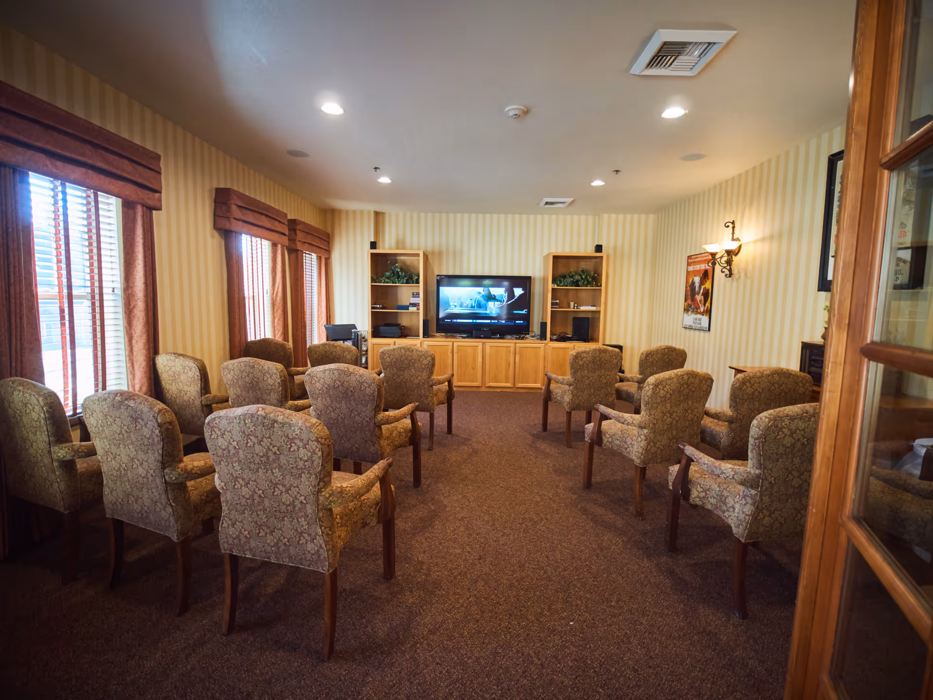 Rows of upholstered chairs facing a television and wooden entertainment center in a cozy communal lounge.