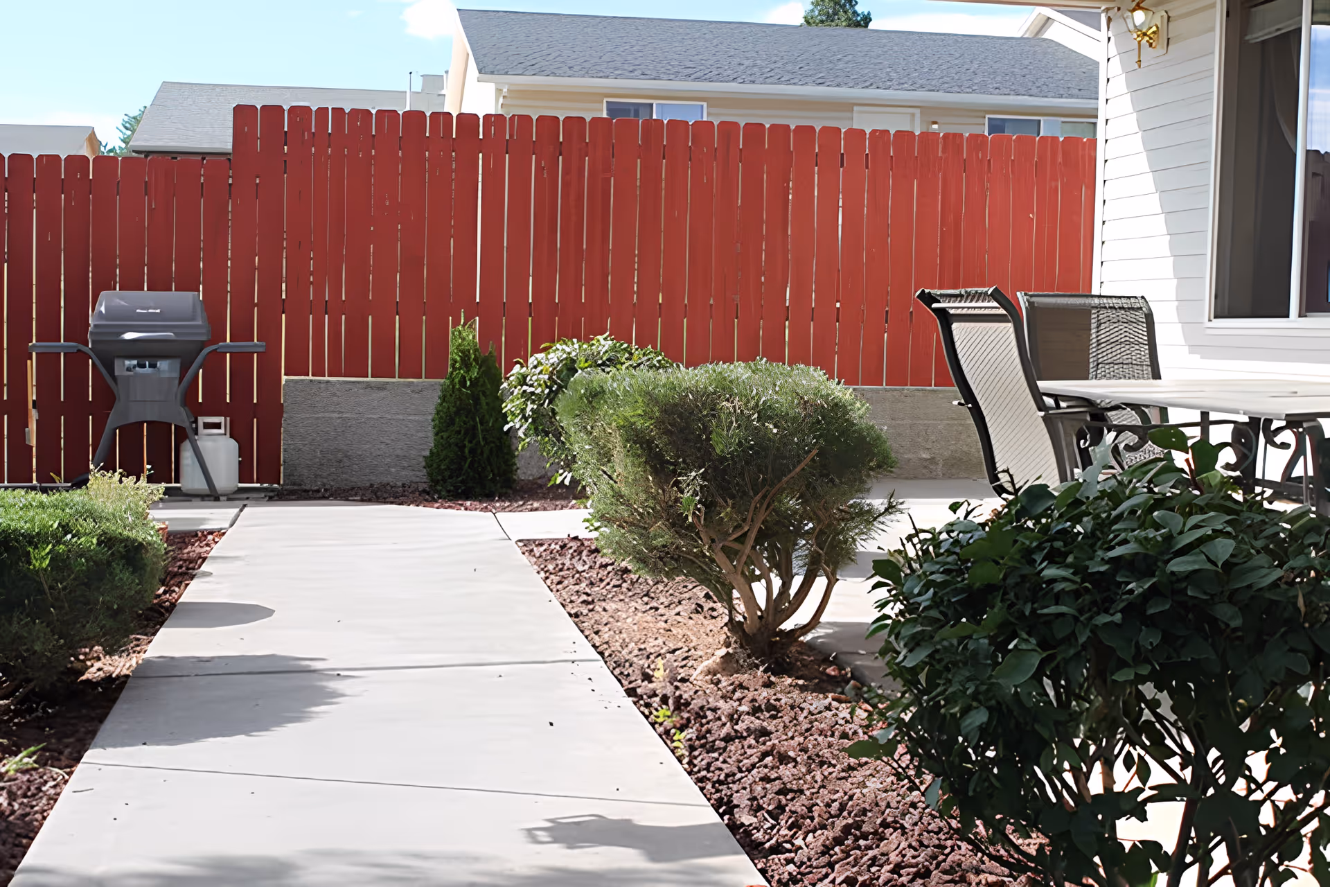 Outdoor patio area with a concrete walkway, bushes, a red wooden fence, a barbecue grill, and a table with chairs next to a white building.
