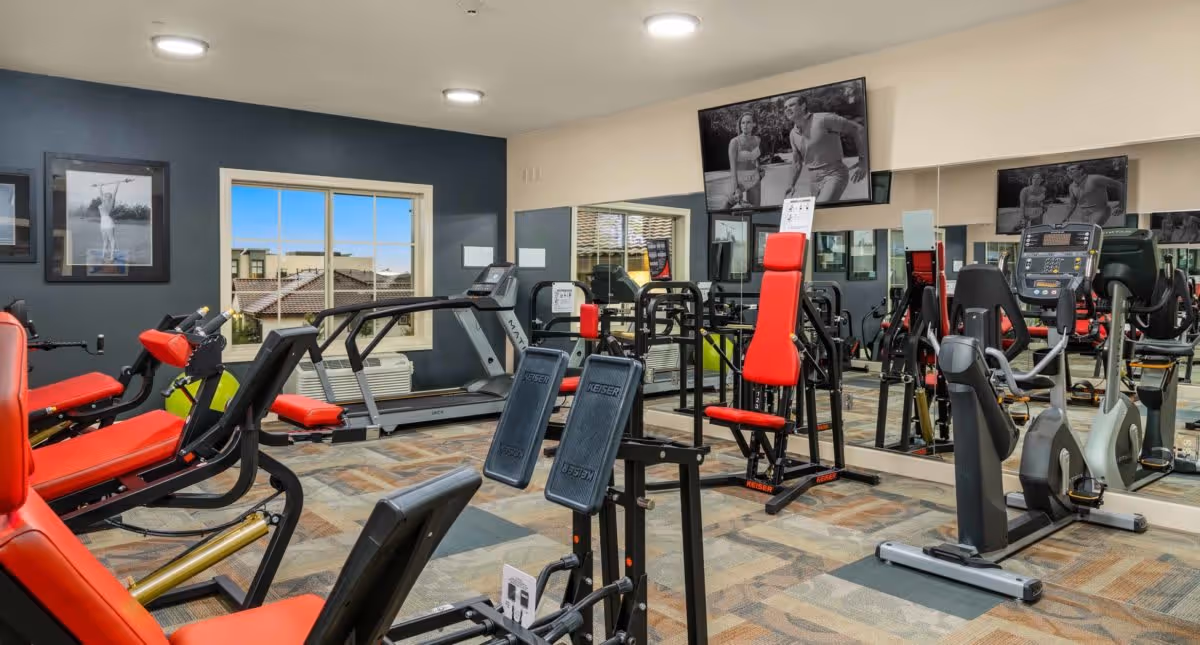 A fitness room with various exercise equipment including treadmills, stationary bikes, and weight machines with red padded seats. The room has a large mirror on one wall, a window showing a view of rooftops outside, and black and white framed photos on the walls. A television is mounted above the mirror displaying a black and white image of a man and woman exercising.