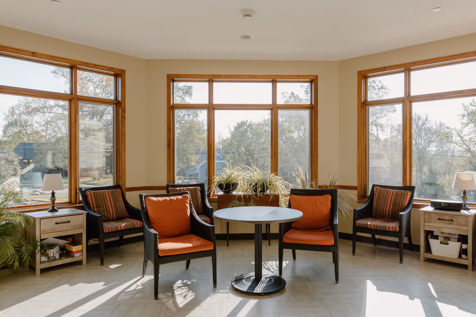 A bright sitting area with large windows letting in natural light. The room has five black wicker chairs with orange and striped cushions arranged around a small round table. There are two wooden side tables with lamps and plants placed on and around them. Outside the windows, trees and a clear sky are visible.