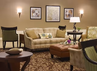A cozy living room area with a beige sofa adorned with decorative pillows, a brown tufted ottoman, several upholstered chairs, a round side table with a lamp and plant, and framed artwork on the wall above the sofa. The room has warm lighting from wall sconces and a patterned carpet.