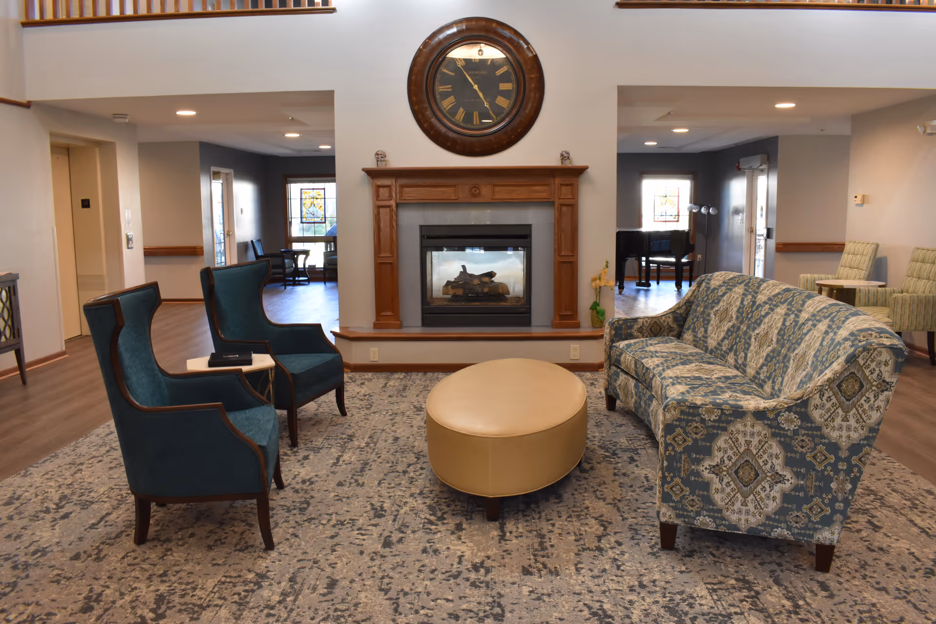 A cozy living room area in a senior living facility featuring a patterned blue and beige sofa, two teal armchairs, and a beige oval ottoman on a large area rug. A wooden fireplace with a glass front is centered on the far wall, above which hangs a large round clock with Roman numerals. The room has wood flooring and neutral-colored walls, with additional seating and a piano visible in the background.